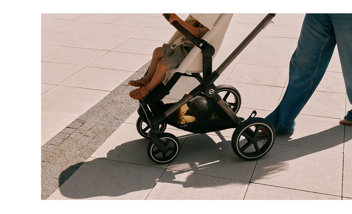 A toddler sits in the CYBEX EOS LUX stroller, photographed from the side on a sunny summer day. The stroller is cropped into the frame from above.