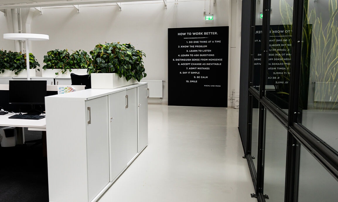 A modern office with white cabinets, computers, and large green plants. A black sign on the wall displays tips for improving work methods.