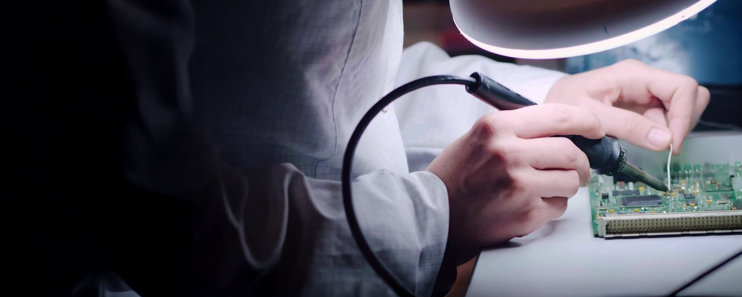 A technician soldering a circuit board under a magnifying lamp.