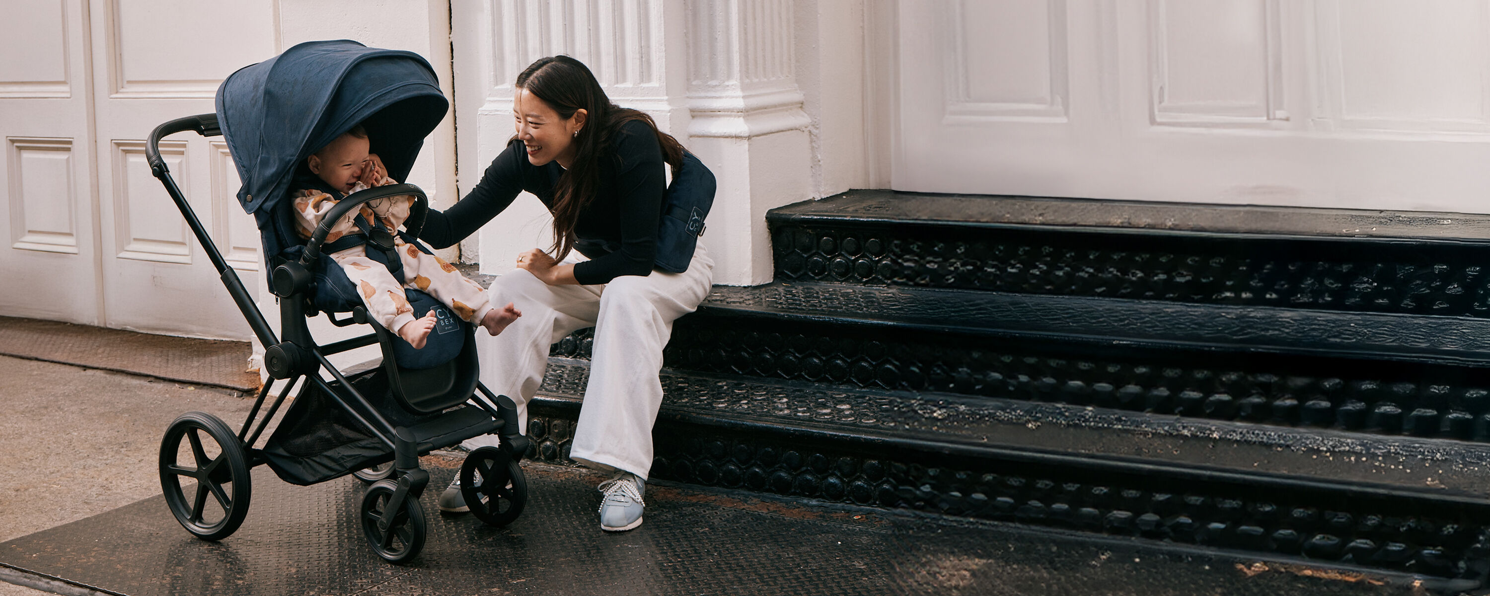 A woman sitting on a flight of stairs, smiling at her baby inside a CYBEX stroller.