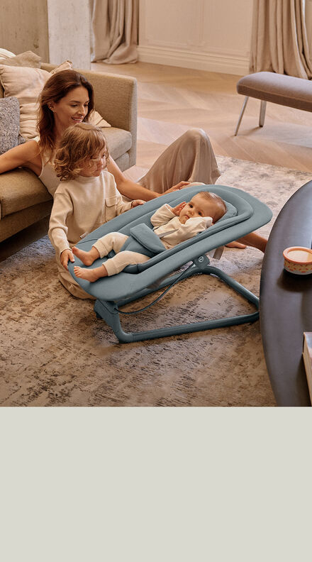A mother sitting on a floor, resting against a couch as her children are sitting next to her. Her baby is in a Lemo Bouncer.