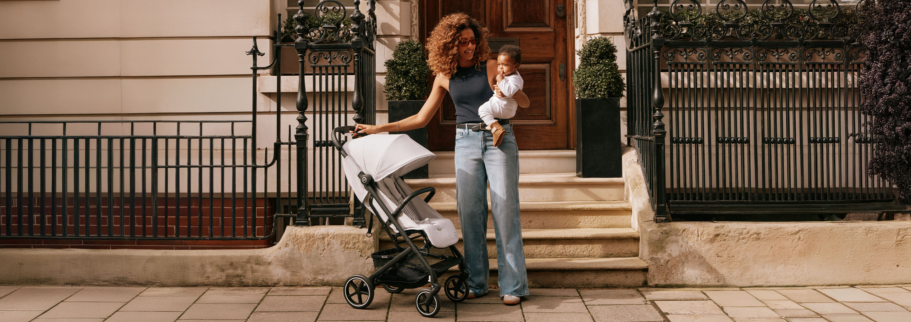 A mother stands with a baby in her arms next to a Beezy lightweight compact stroller on steps in front of a building.