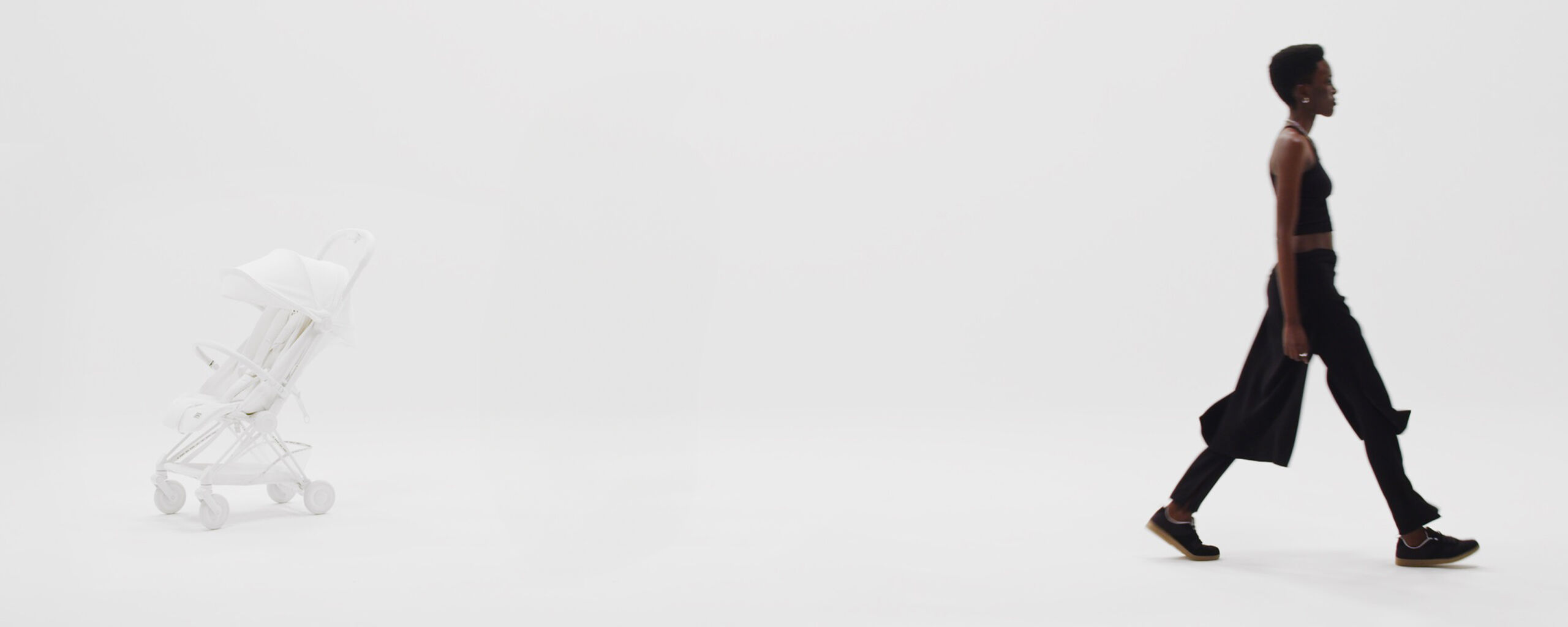 A white stroller is set against a white background, while a person in black clothing walks from right to left.