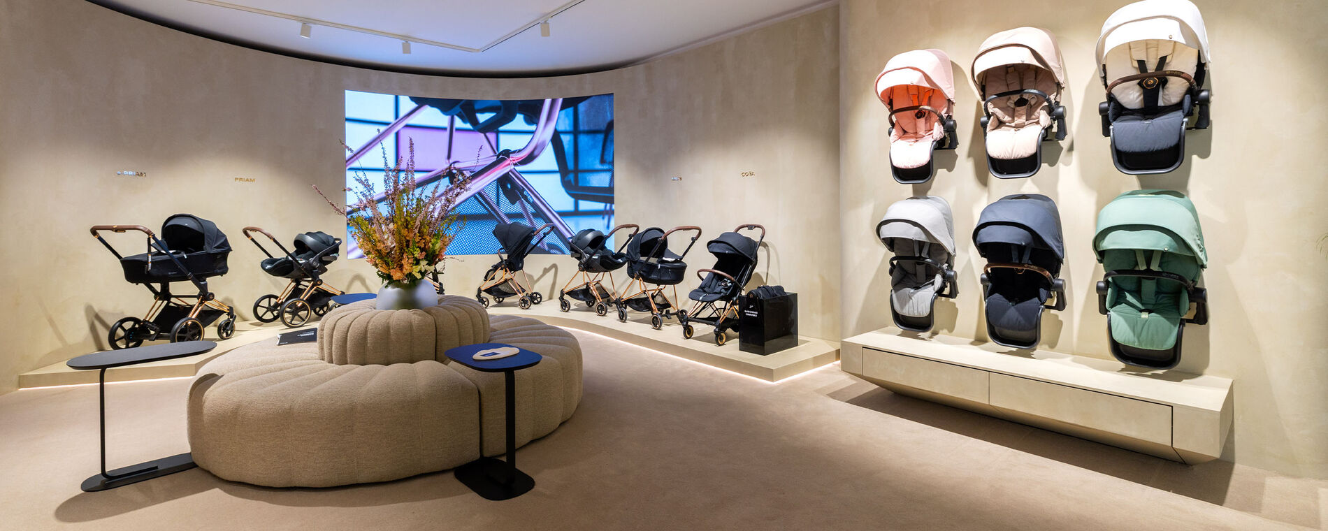 CYBEX store interior with a round beige sofa and two round black coffee tables. Behind it, CYBEX Platinum pushchairs with different attachments are displayed, along the wall seat packs in various colours.