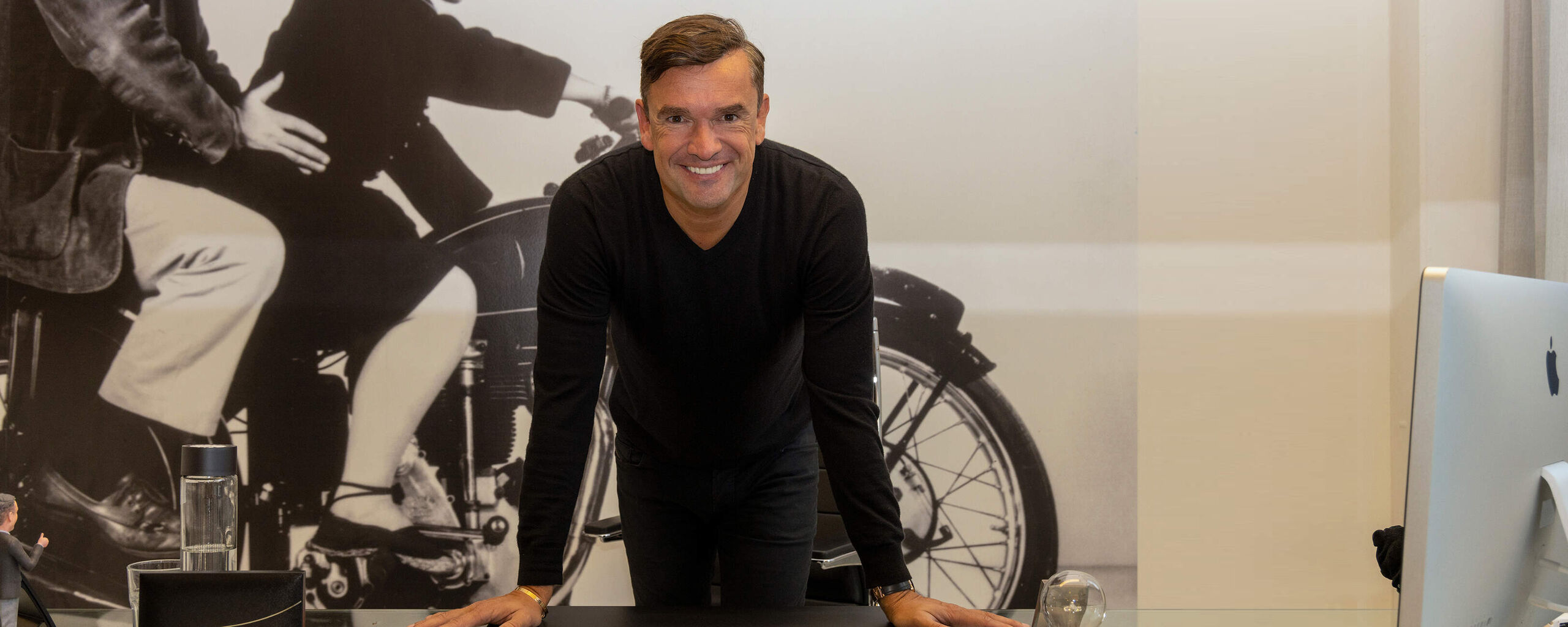 A man leaning over a table, smiling at the camera, with a black-and-white photo of a motorcycle in the background.