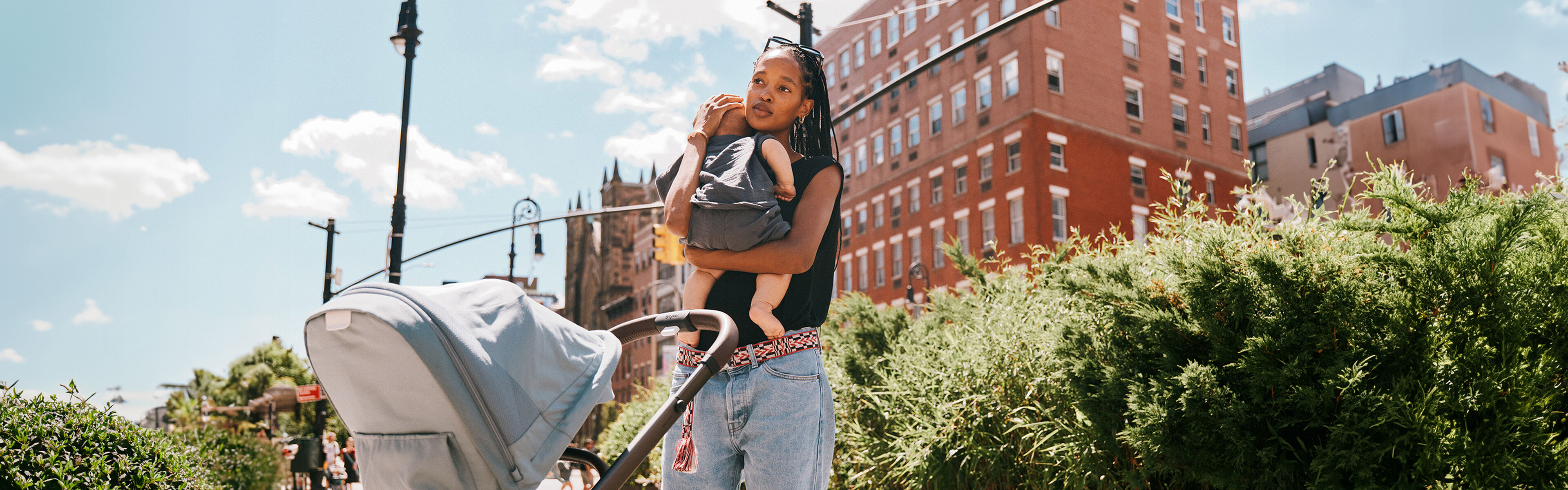 A woman carrying a child and pushing a pram on a sunny pavement, surrounded by green bushes and red brick buildings.