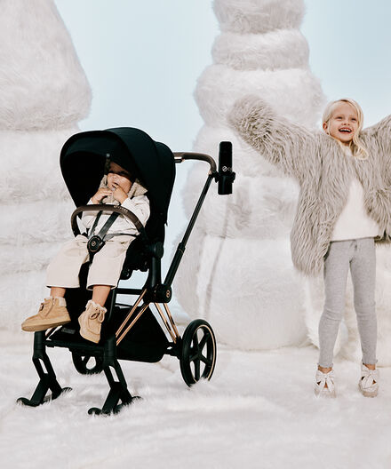 Un jeune garçon assis dans une poussette CYBEX Priam avec des skis. À sa droite, une fille sourit en levant joyeusement les bras. 