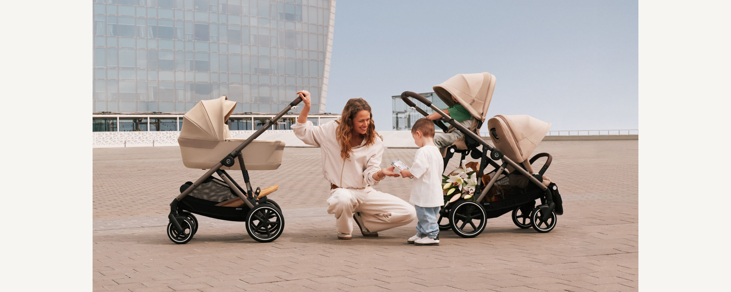 A mother is kneeling on the ground next to her child, grinning at him. She has two Gazelle S strollers next to her, one in single mode, one in sibling mode.