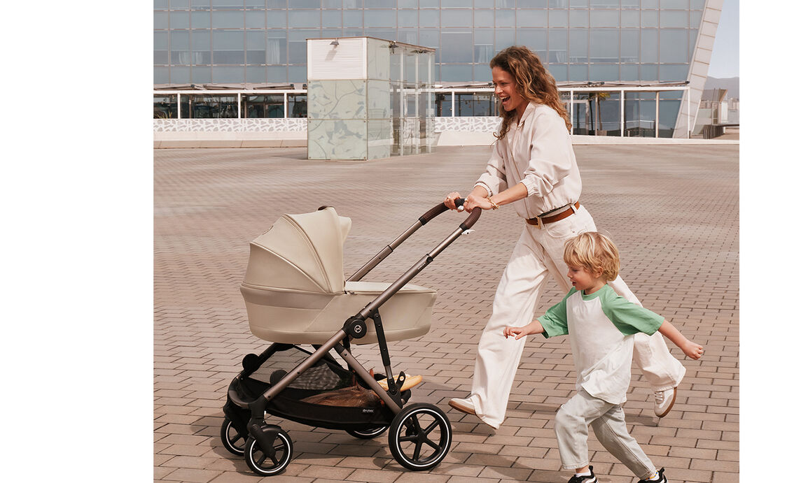 A woman pushing her Gazelle S stroller with a Cot, laughing as she is running together with the son next to her.
