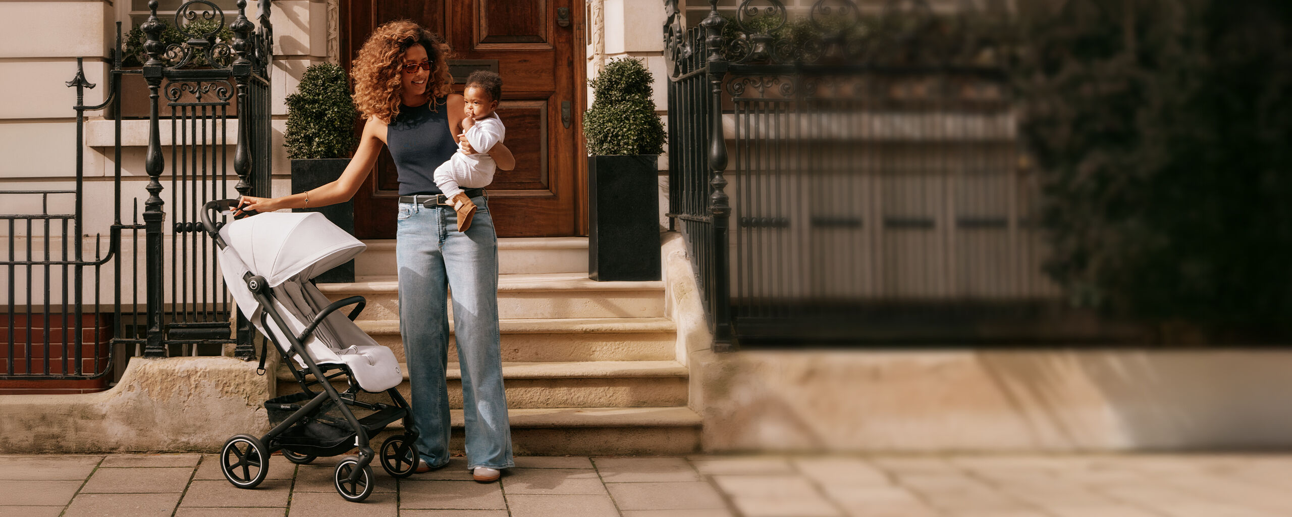 A woman stands next to a Beezy lightweight compact stroller on a staircase, holding a baby in her arms.