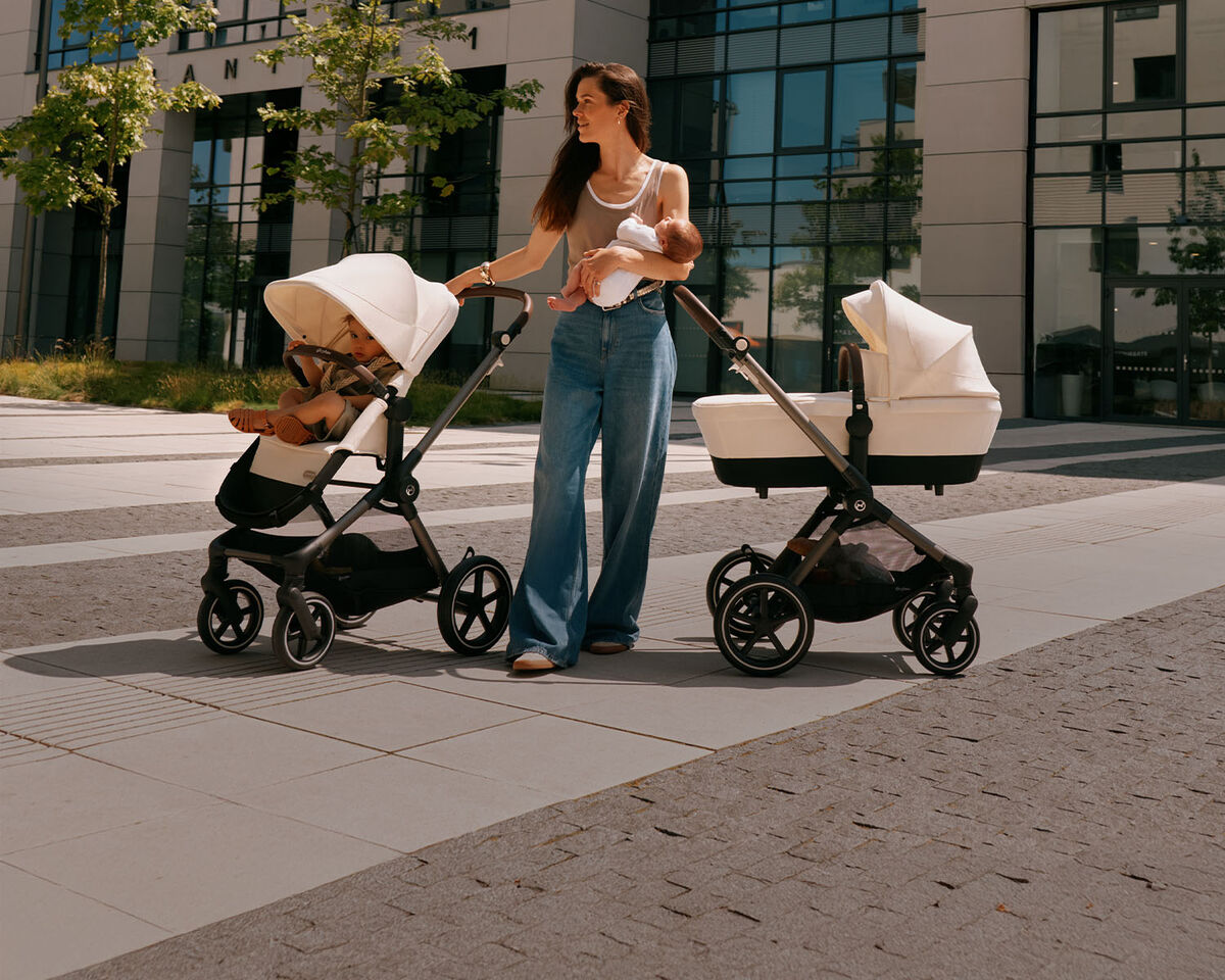 A woman holding her newborn baby against her chest, two Eos Lux strollers next to her.
