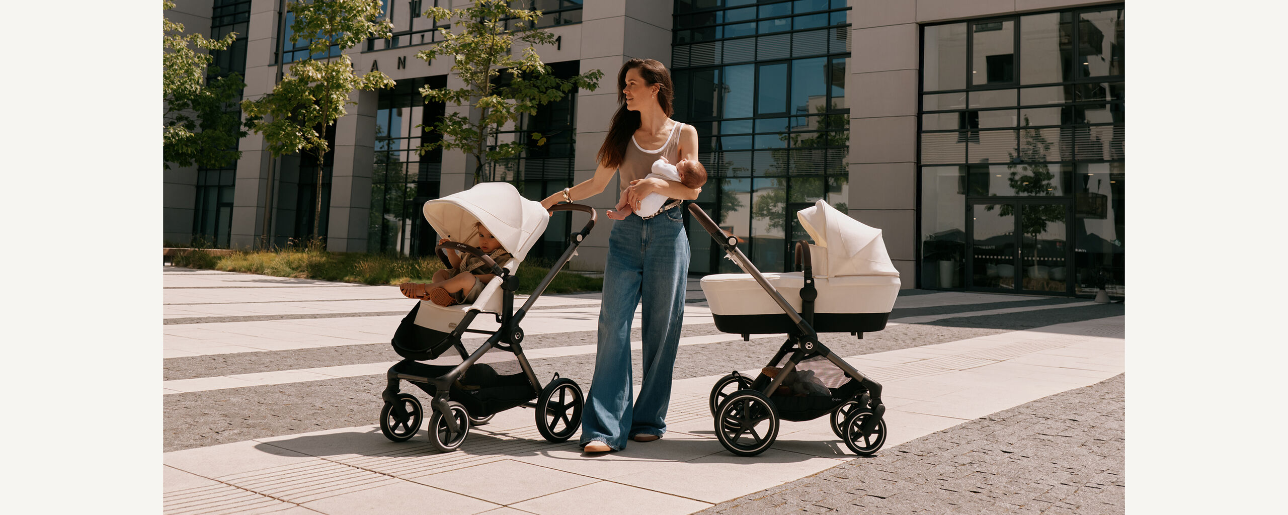 CYBEX stroller duo &ndash; mother holding baby beside two strollers outdoors, one with toddler seat, one with carrycot, in sunny urban scenery
