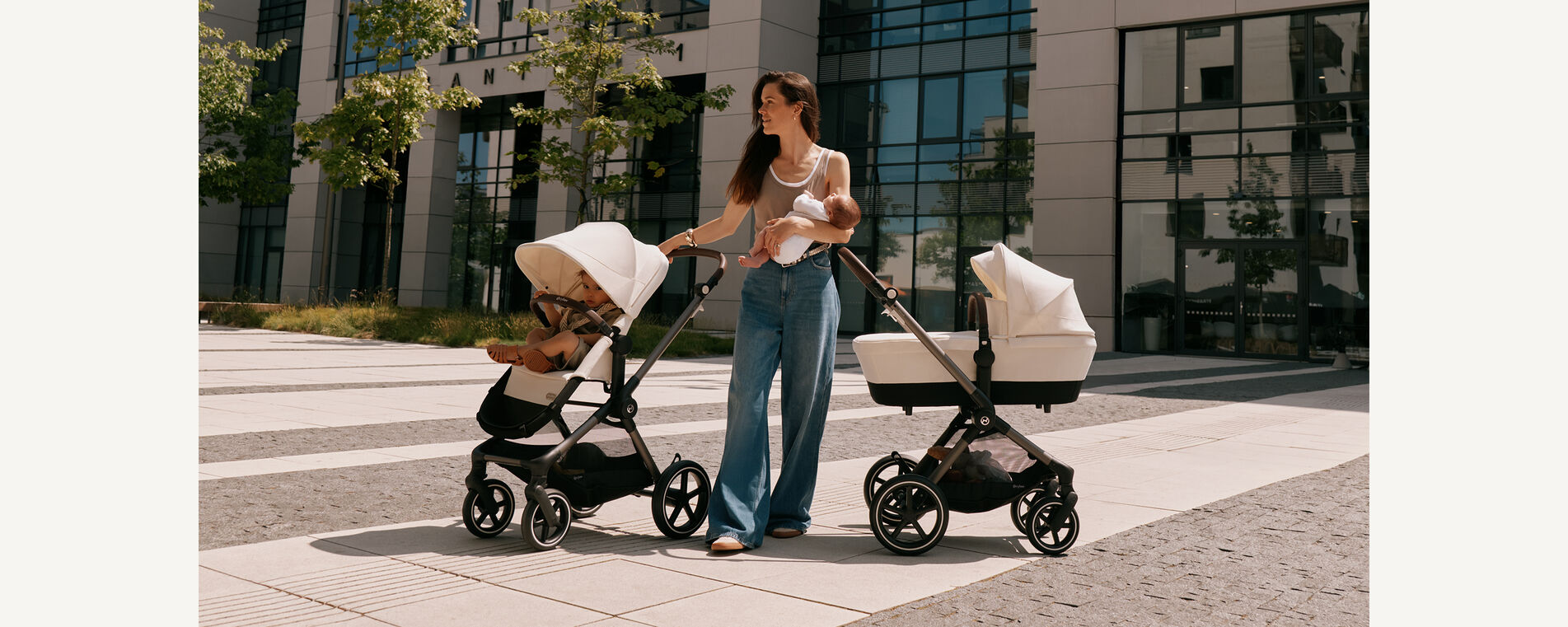 CYBEX stroller duo – mother holding baby beside two strollers outdoors, one with toddler seat, one with carrycot, in sunny urban scenery