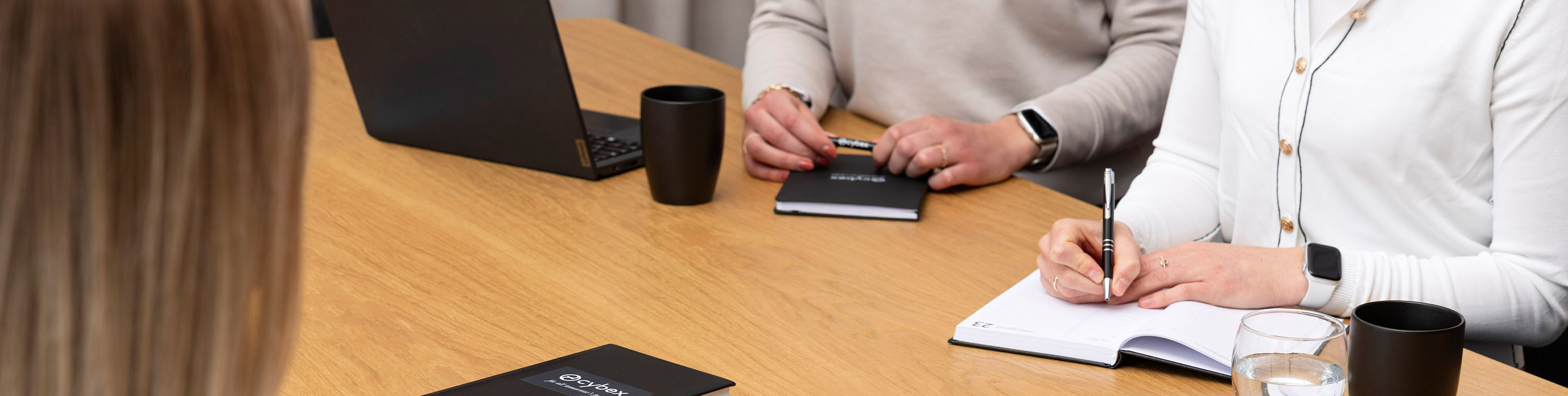 People sitting at a meeting table, notebooks and pens on the table, laptop and coffee cup visible.
