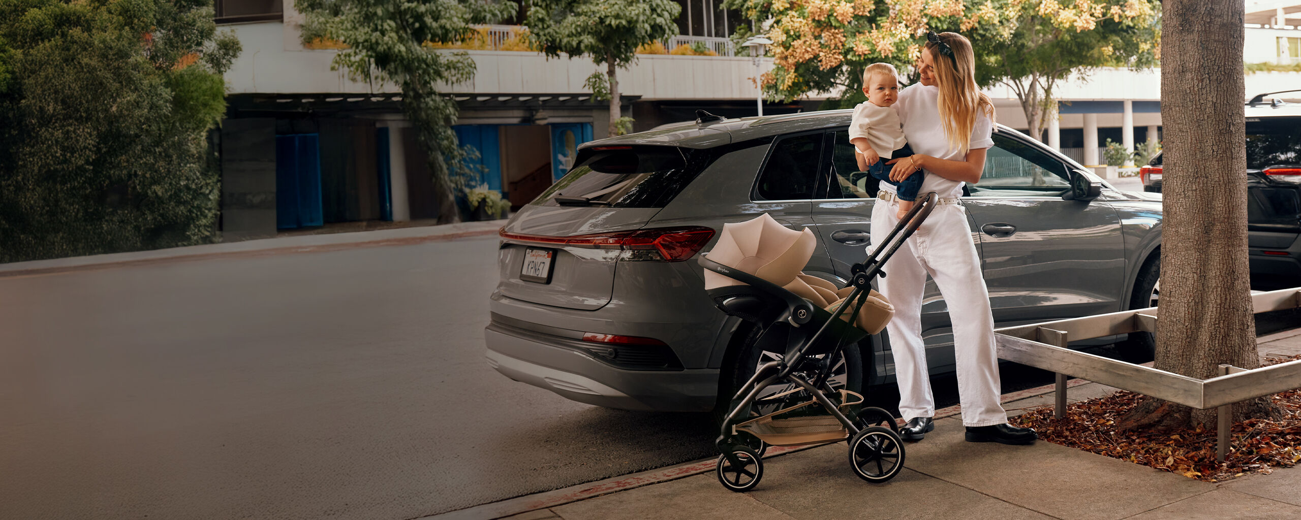 A mother standing before a car, holding her child in her arms and an Eezy S Twist +2 buggy with a Cloud G attached is before her.