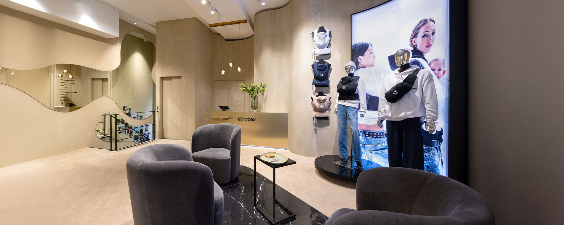 CYBEX store interior with two anthracite armchairs and a coffee table. A black shelving unit with reflections and elegant pendant lights decorates the wall. The Urban Mobility collection carriers are displayed alongside a poster.