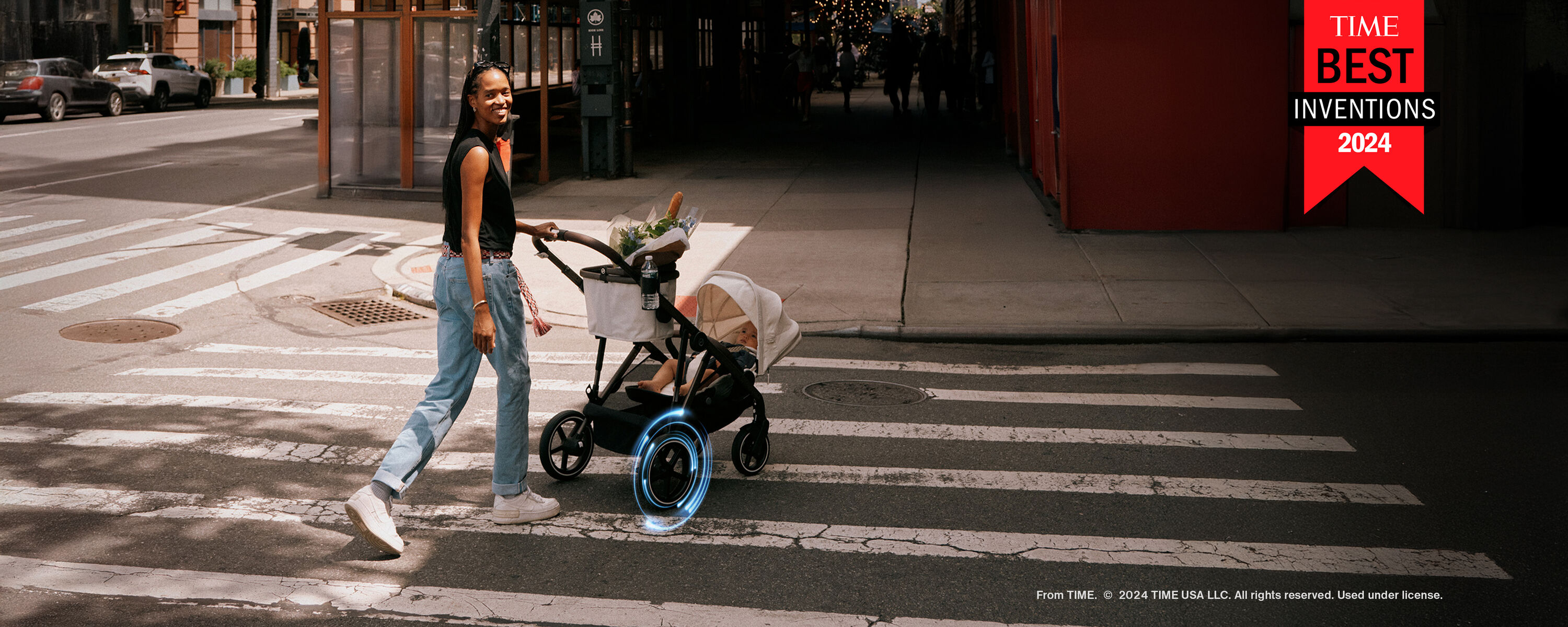 Een vrouw met lang haar steekt met een Kinderwagen een zebrapad over in een stad, omringd door moderne gebouwen.