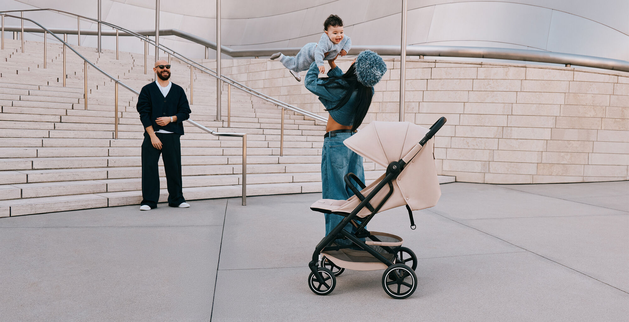 A woman playing with her son by throwing him up into the air while the father watches, laughing. A Beezy buggy stands in front of them.