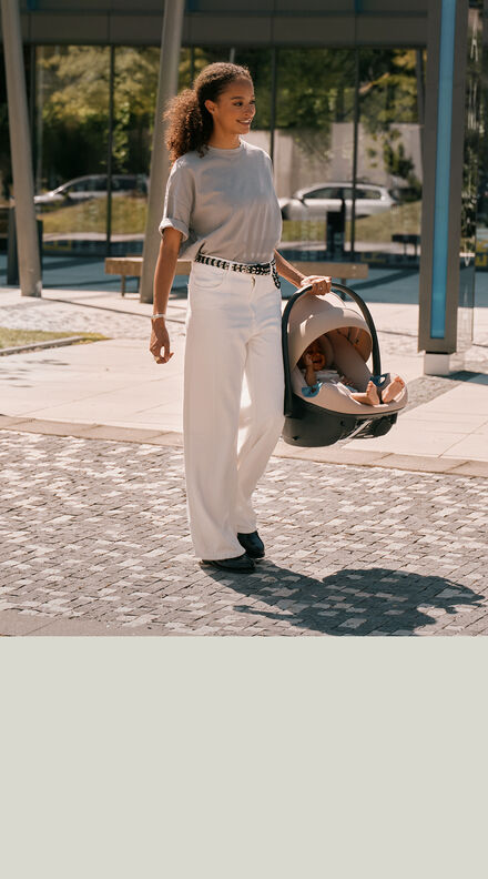 Woman carrying the CYBEX Aton G2 infant car seat in beige with a baby seated inside, walking along a sunlit pathway.