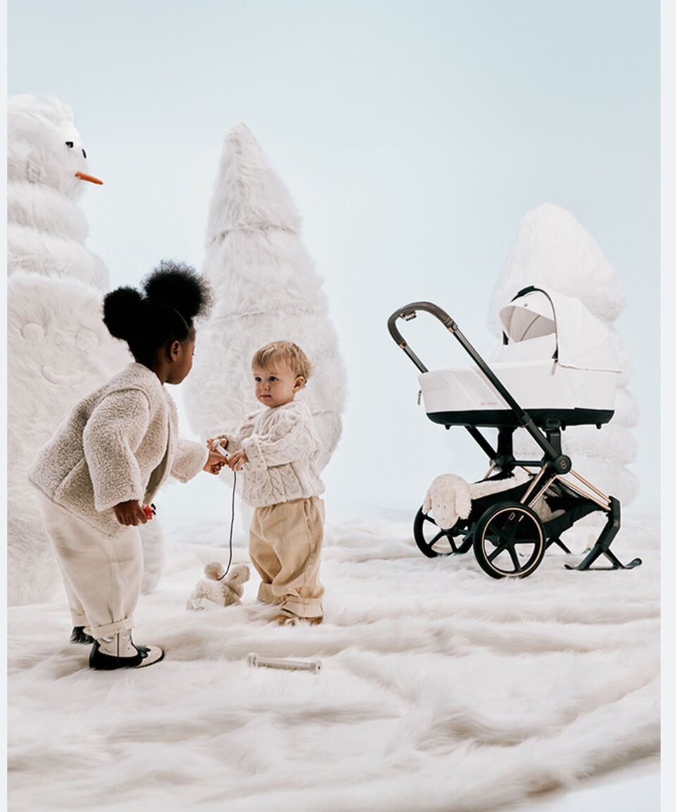Une petite fille et un garçon jouant avec un jouet. À l’arrière-plan, on aperçoit des sapins de Noël blancs et une Priam équipée de skis.