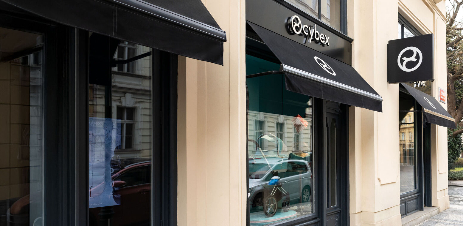 Three large display windows of the CYBEX store with elegant logo awnings. The glass reflects the historic old town façades, creating a striking contrast with the modern store design.