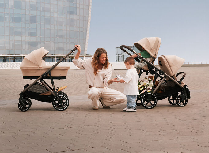 A woman kneeling on the ground, handing something to her child. On the left side, a Gazelle S stroller with a Cot, on the right, a Gazelle S with two seat units.