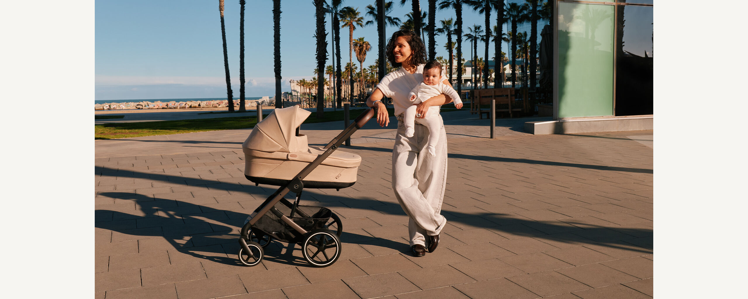 A woman leaning casually against her Balios S Lux stroller with a grin, holding her baby in her left arm.