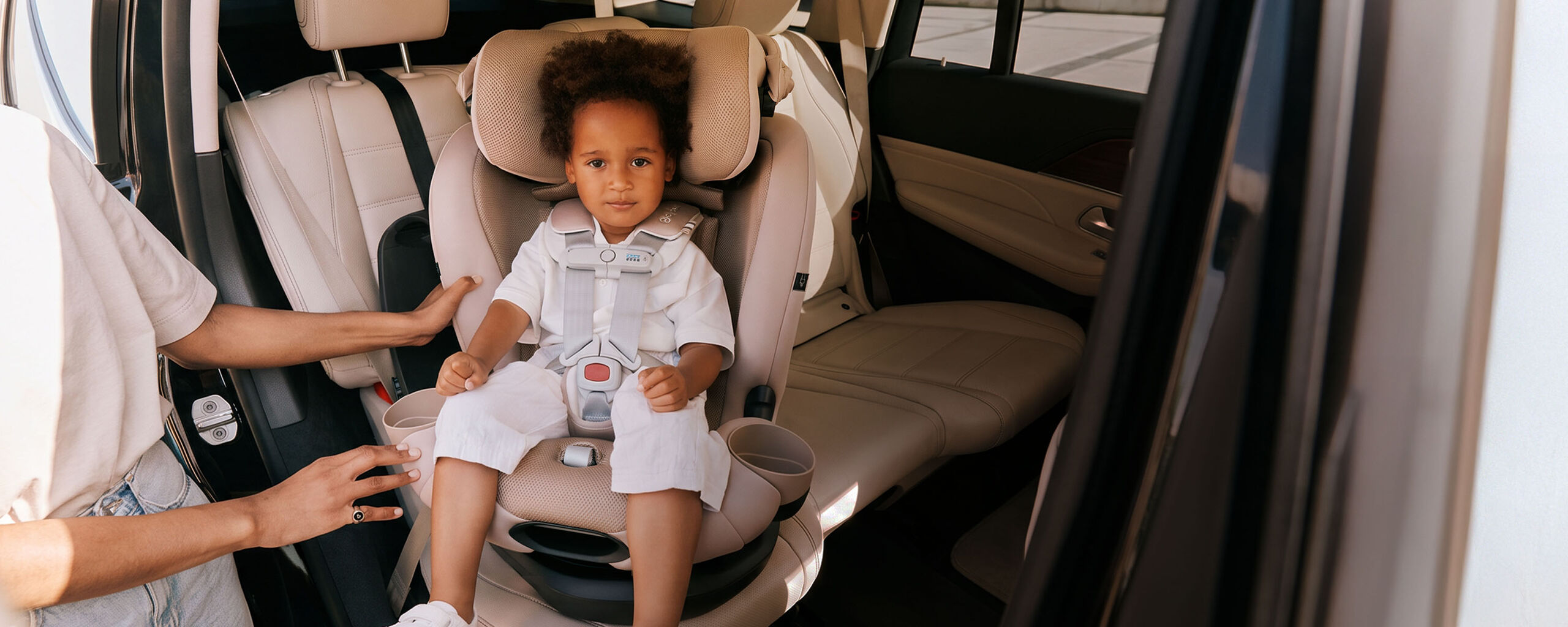 A child in the back of a car, sitting in a car seat that is turned towards the open car seat door that his mother stands at.