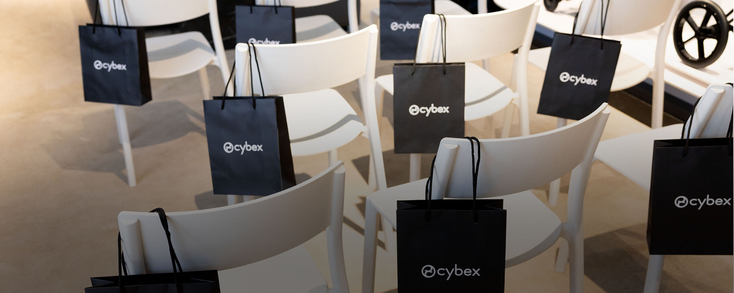 Blonde woman with a doll in a CYBEX baby carrier, explaining something while looking down. Audience members are visible only from the back, with CYBEX Priam campaign displays in the background.