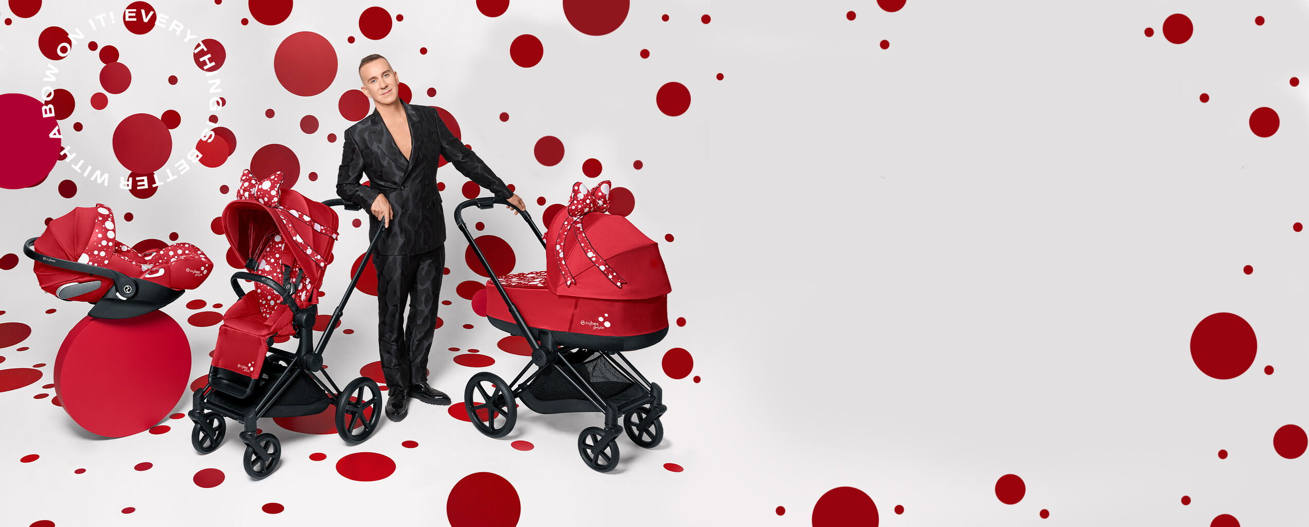 A man in a black suit stands next to three red prams with white polka dots, against a background of red dots.