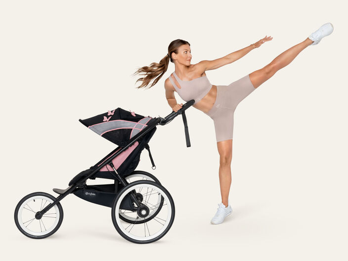 Frau in sportlicher Kleidung macht einen hohen Kick neben einem Kinderwagen mit Schmetterlingsdesign.