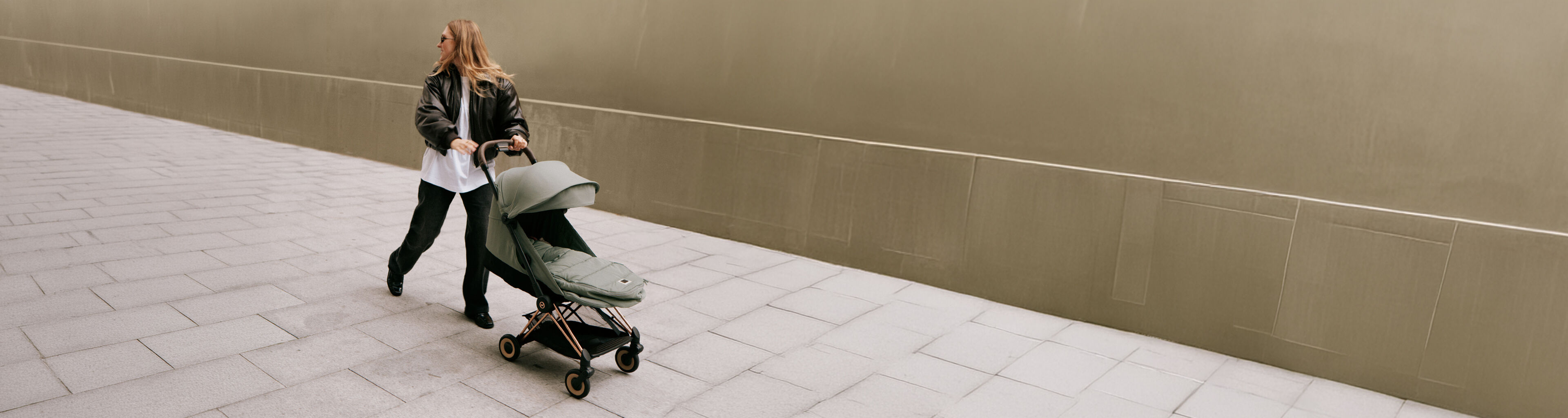 A woman pushes a CYBEX Club pram with a baby inside along a cobblestone path in front of a large building.
