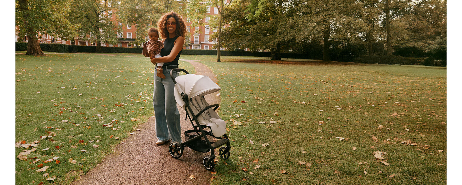 A woman standing in a park next to her CYBEX buggy, holding her child in her arms and laughing into the camera.