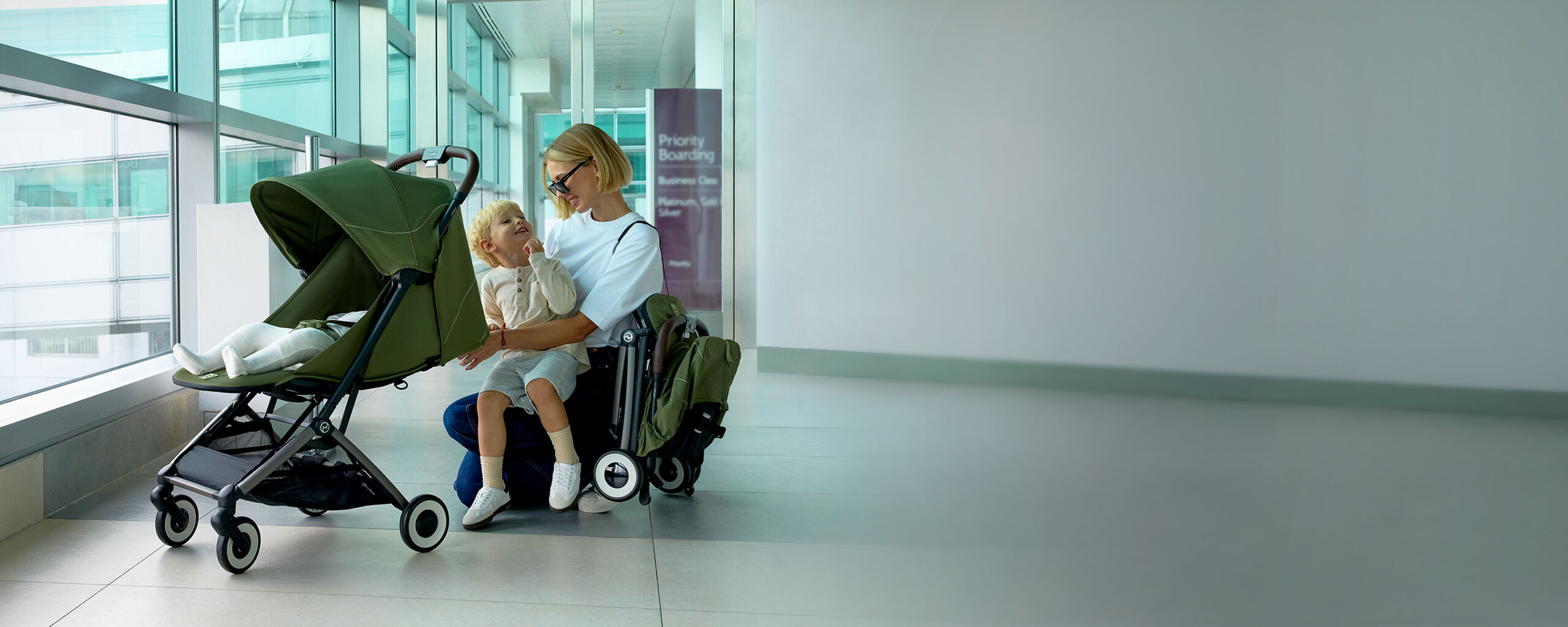 W&oacute;zek podr&oacute;żny Orfeo w nowoczesnym lotnisku, matka siedzi na podłodze z dzieckiem, podczas gdy w&oacute;zek stoi obok nich.