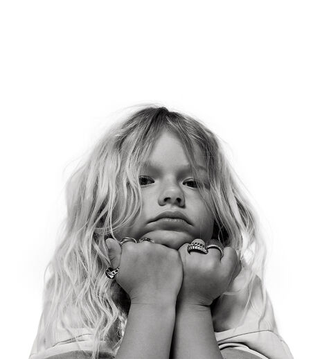 A girl with blonde hair looking into the camera. Her chin is resting on both of her fists that she wears several rings on.
