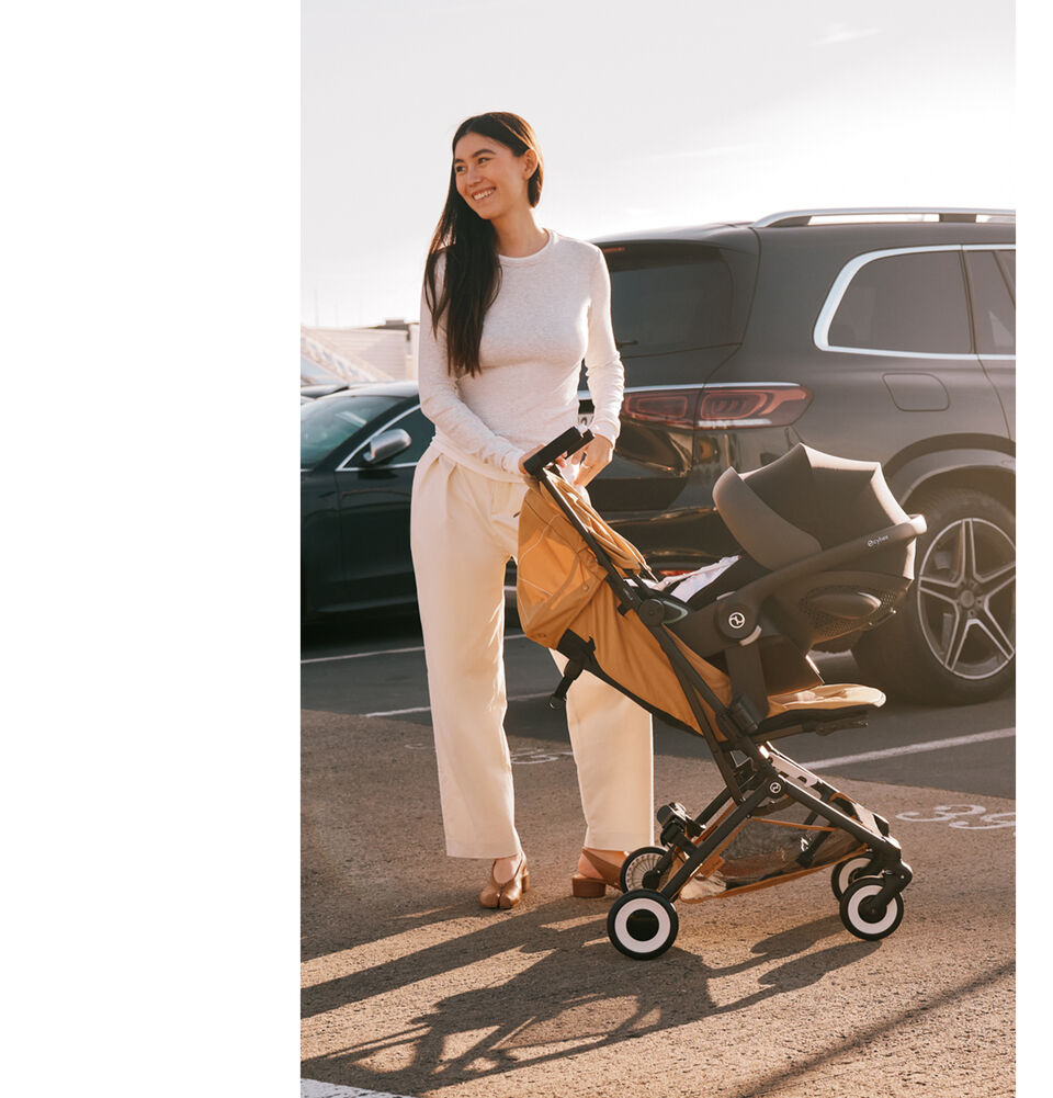 A woman in an outside parking lot in front of two cars. She's holding on to a Libelle buggy with an infant car seat attached in the recline seat pack.