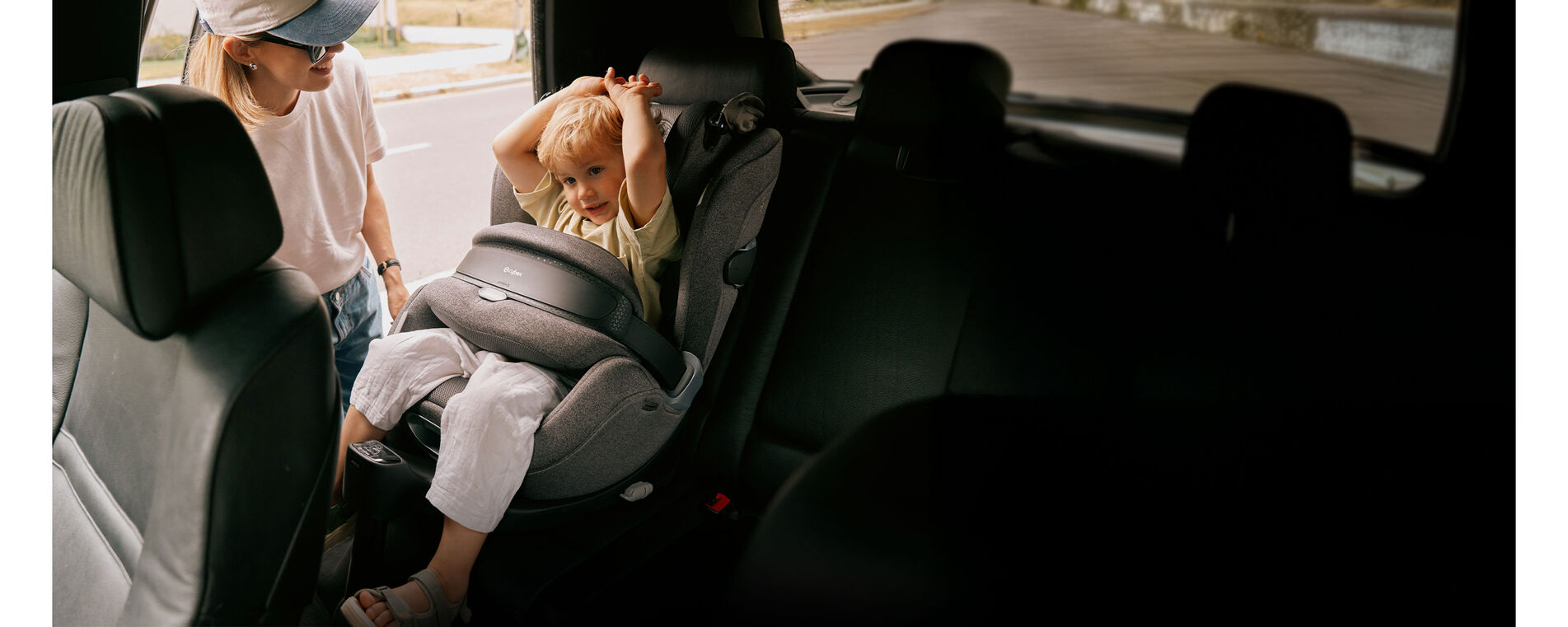 A child sitting inside an Anoris T2 airbag car seat with his hands up. His mother is standing by his side at the open car door and smiling at him.