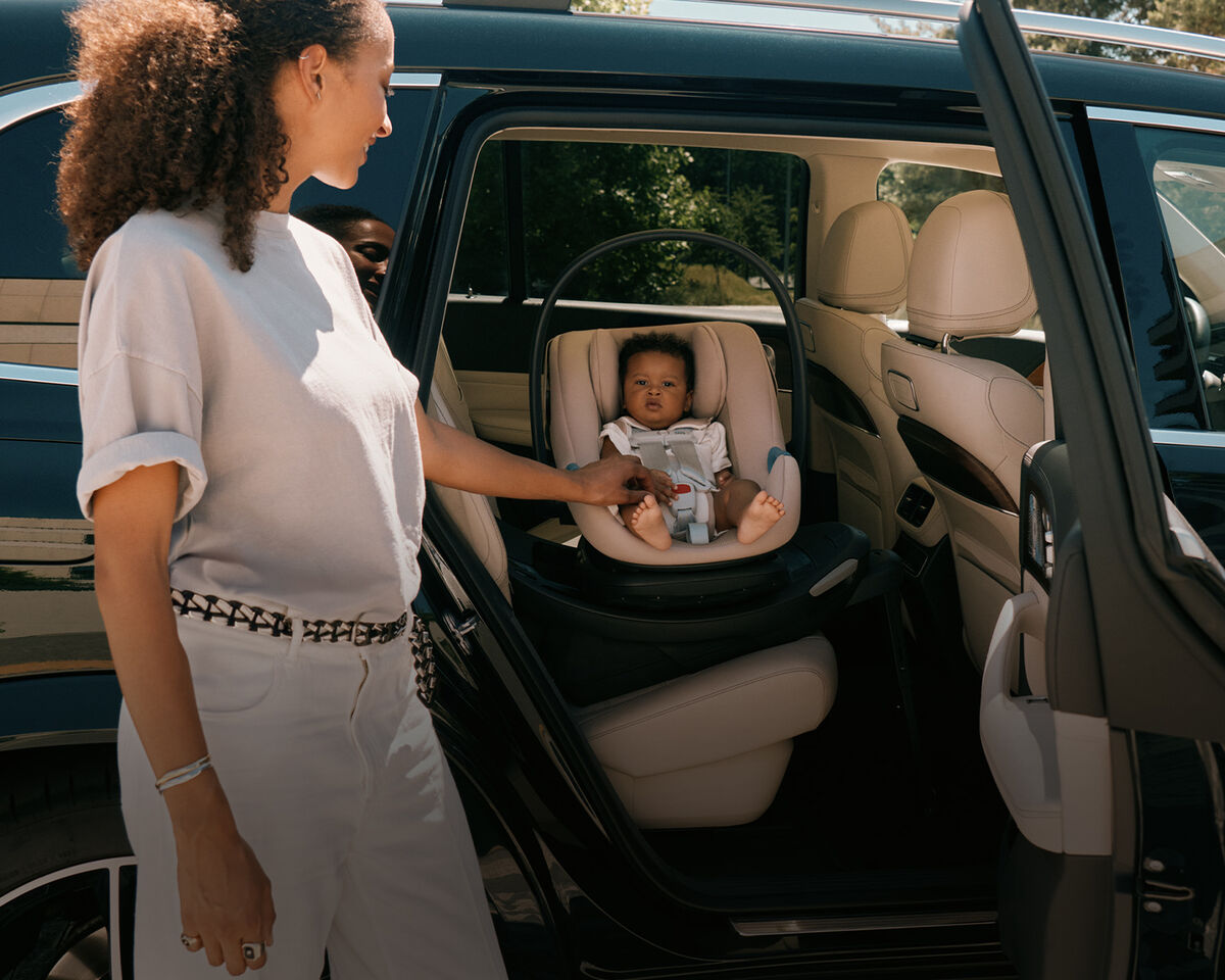A woman standing at an open car door, holding the hand of her baby that's sitting in an Aton G2 Swivel car seat.