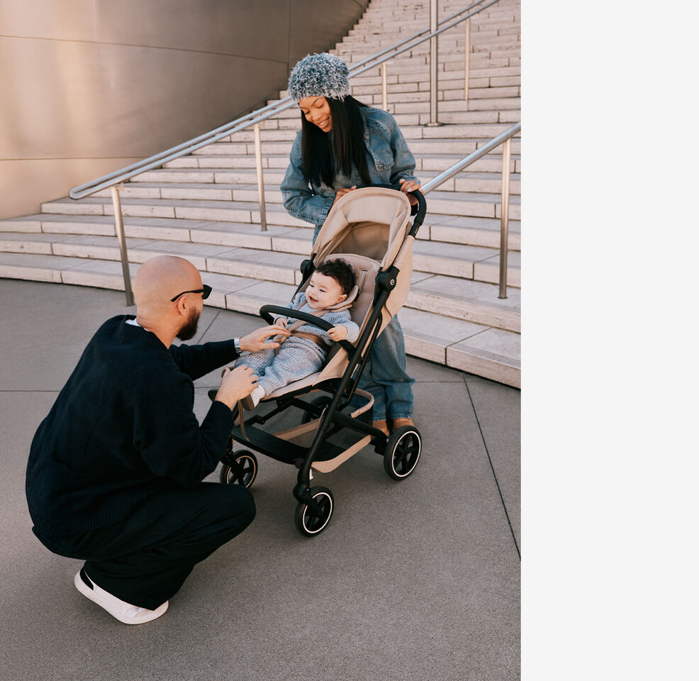 A mother standing behind her Beezy buggy, holding onto the handlebar. Her child is inside, the father kneeling before it and tickling the baby.