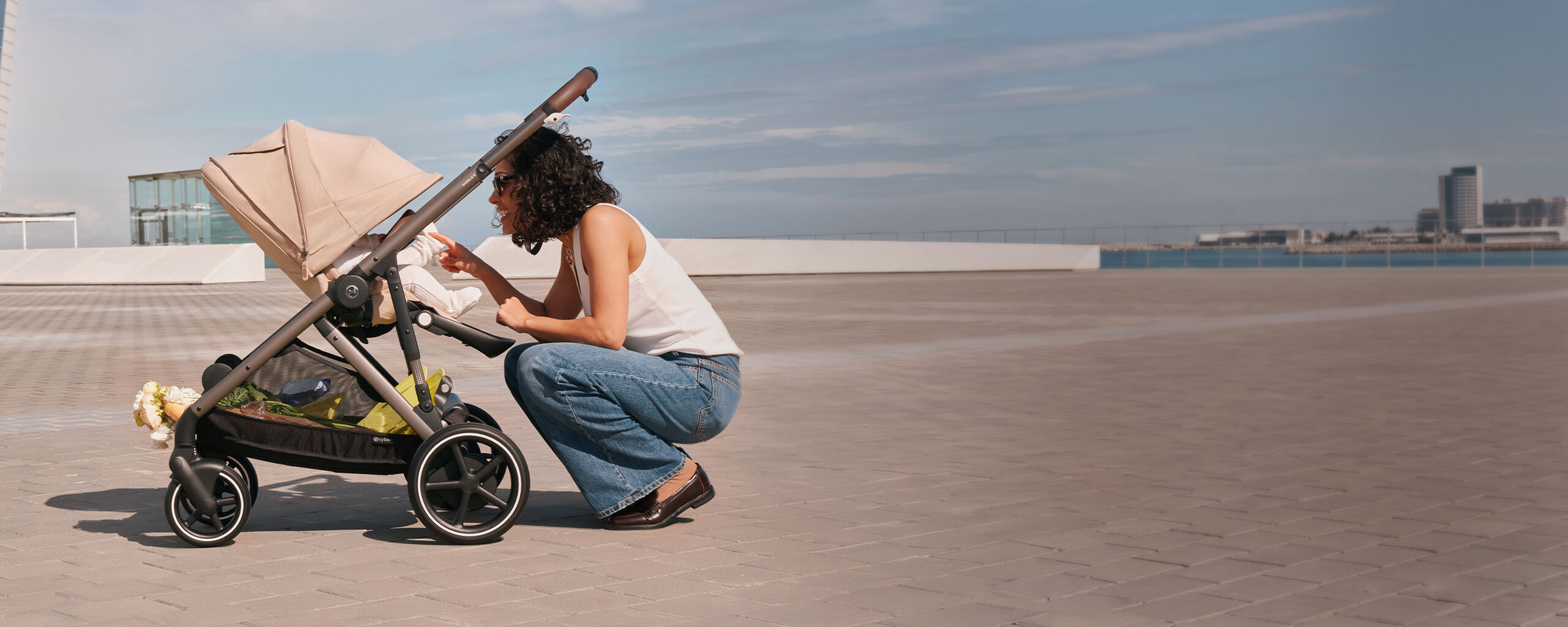 Woman crouching beside the beige CYBEX Gazelle S stroller, smiling and interacting with her baby on a sunlit rooftop terrace.