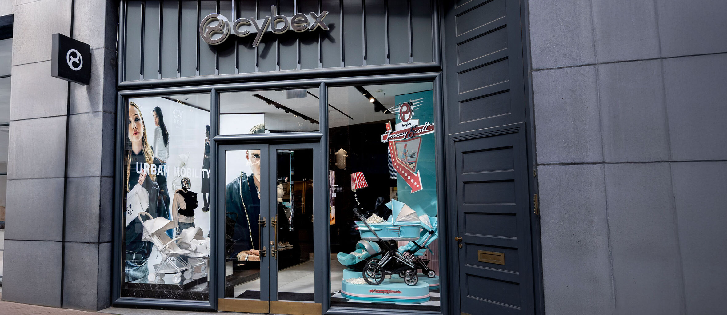 Modern main entrance of the CYBEX store with silver logo sign above the door, framed by anthracite panels. Two shop window displays showcase exclusive CYBEX strollers and car seats.