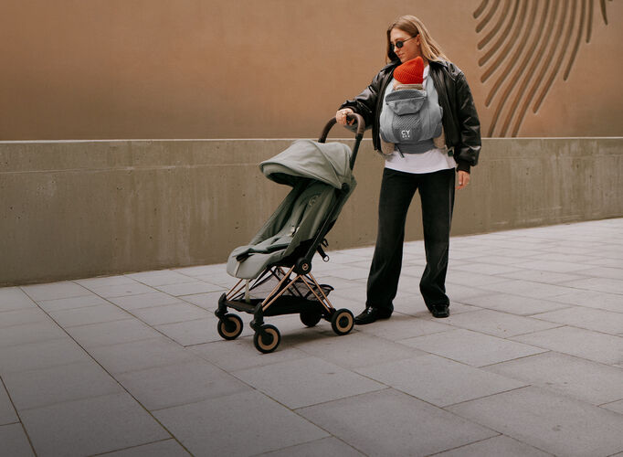 A woman with a baby carrier wearing her baby against her chest, one hand on a Coya lightweight stroller.