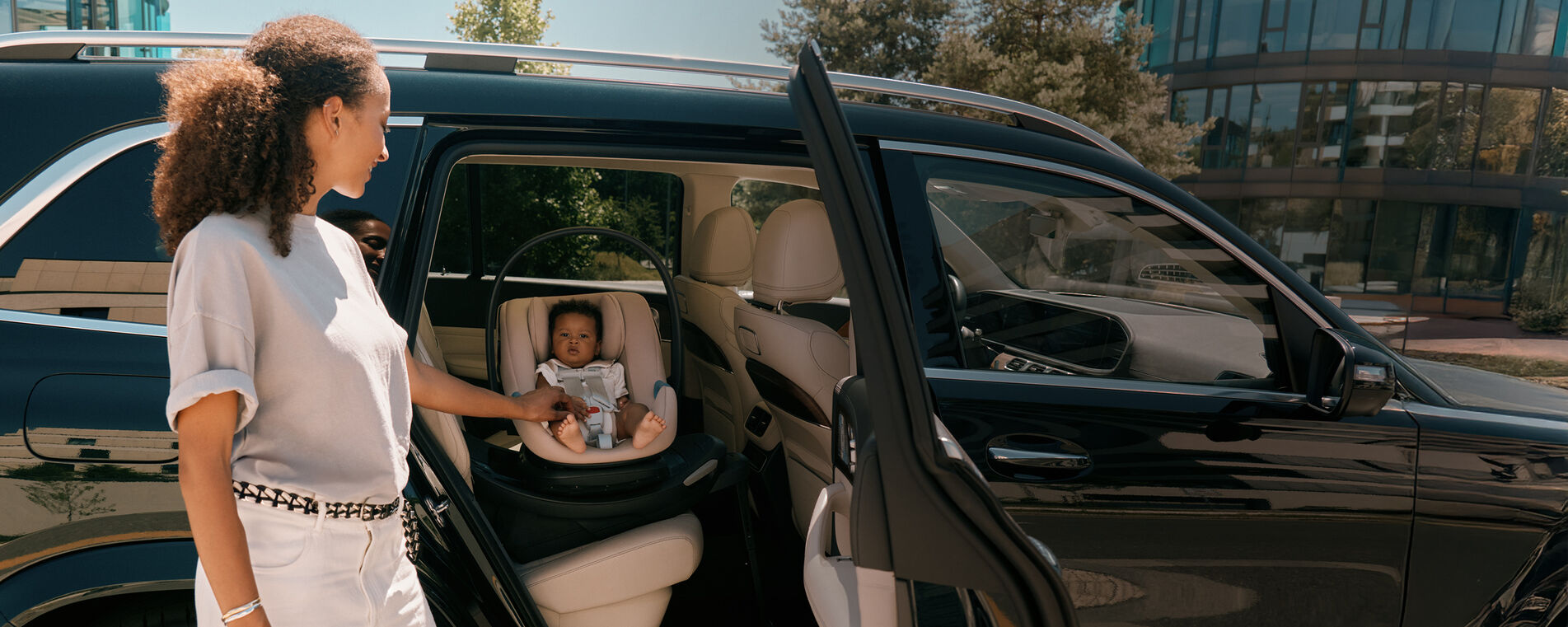 A mother standing at an open car door, holding the hand of her baby that is sitting in an Aton G2 Swivel car seat facing the door.