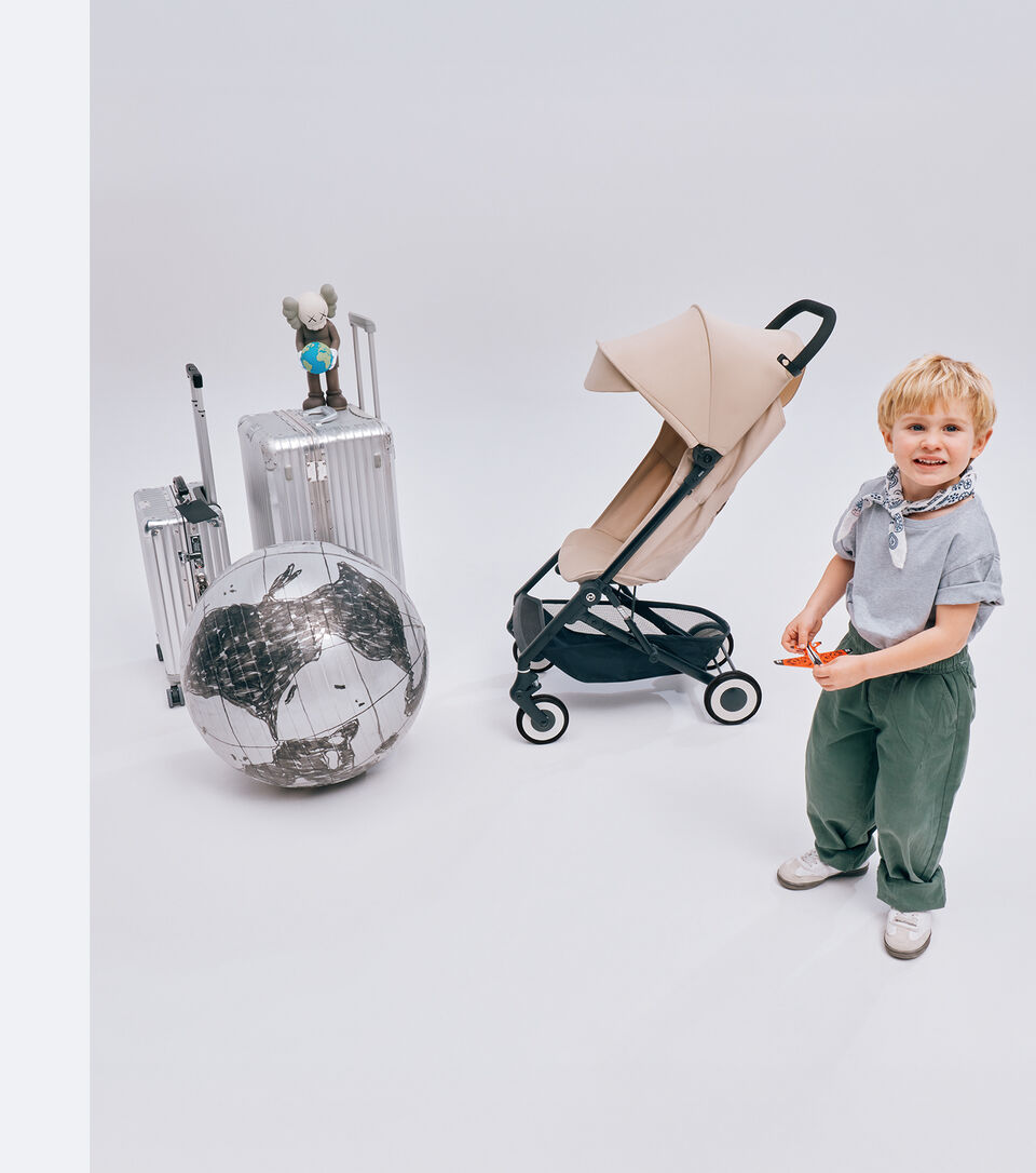 A young boy smiling into the camera, an Agis buggy next to him. Next to that, there are two suitcases and a globe ball in front.