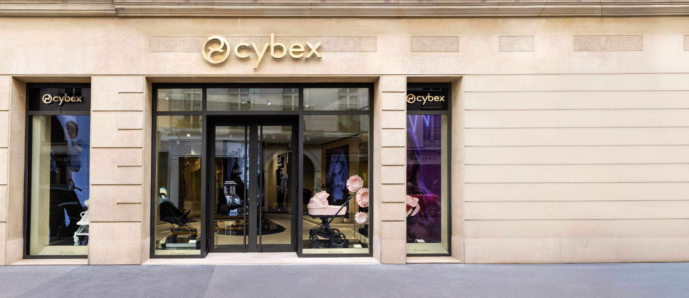 CYBEX store fa&ccedil;ade in beige stone with a shiny gold logo above the main entrance, anthracite window frames, and glass doors. Shop windows display premium strollers, featuring a pink model as the highlight.