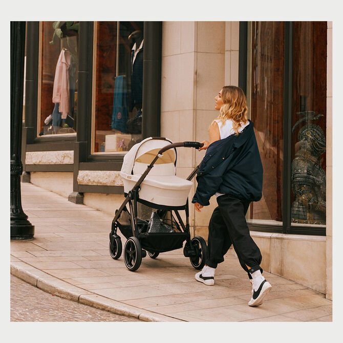 A woman pushing a pram along a pavement in front of a shop window.