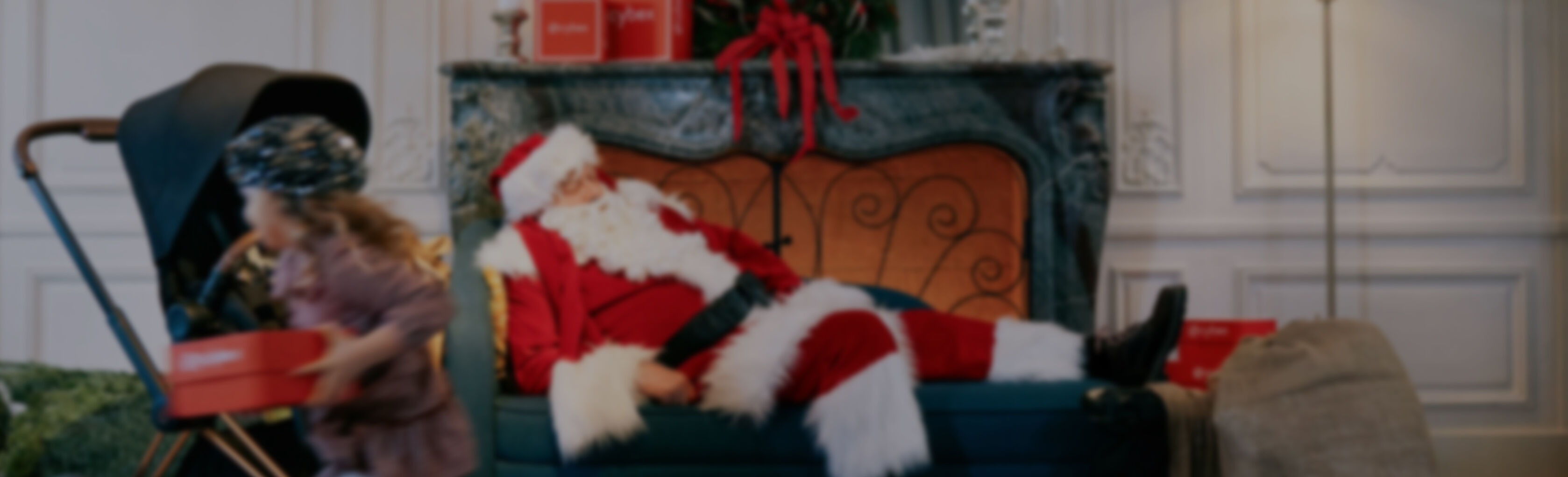 Scena świąteczna ze śpiącym Mikołajem na sofie przed kominkiem, dziecko z pudełkiem prezentowym obok w&oacute;zka