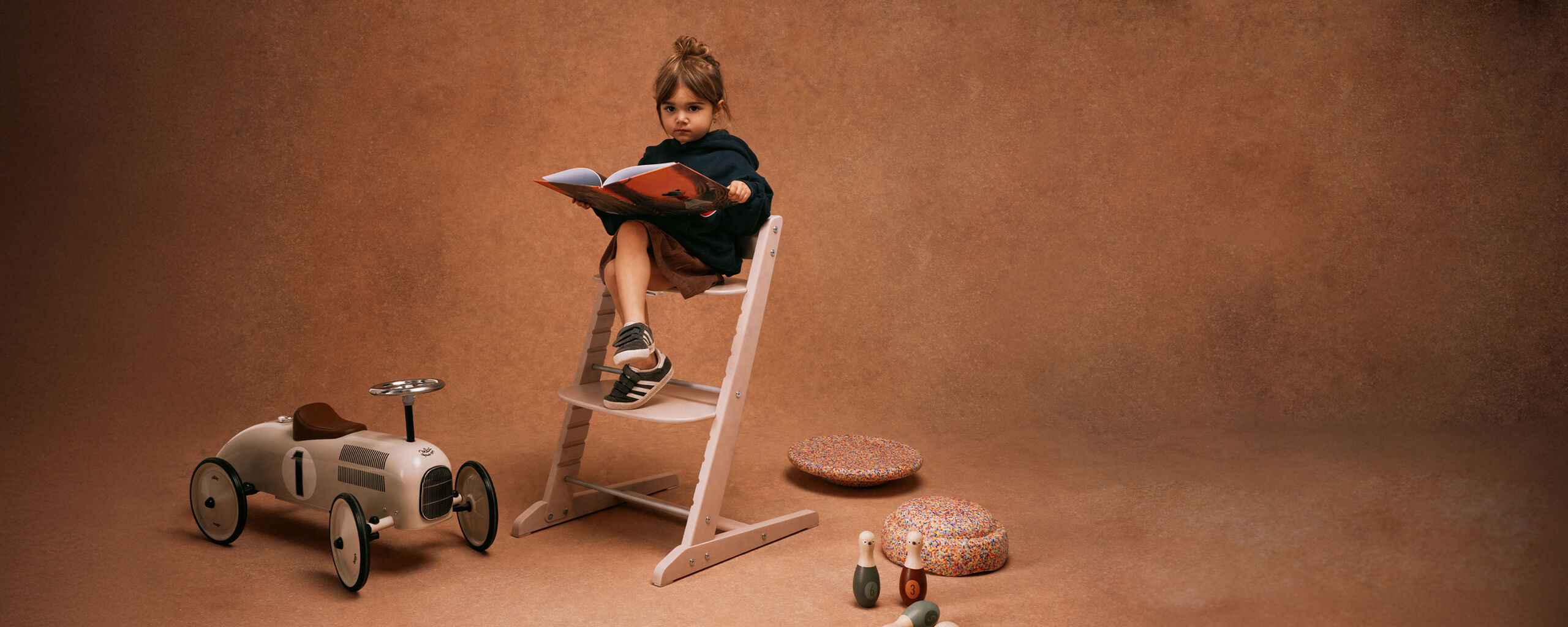 A young girl sitting on an Iris chair, holding a book while toys are scattered around her.