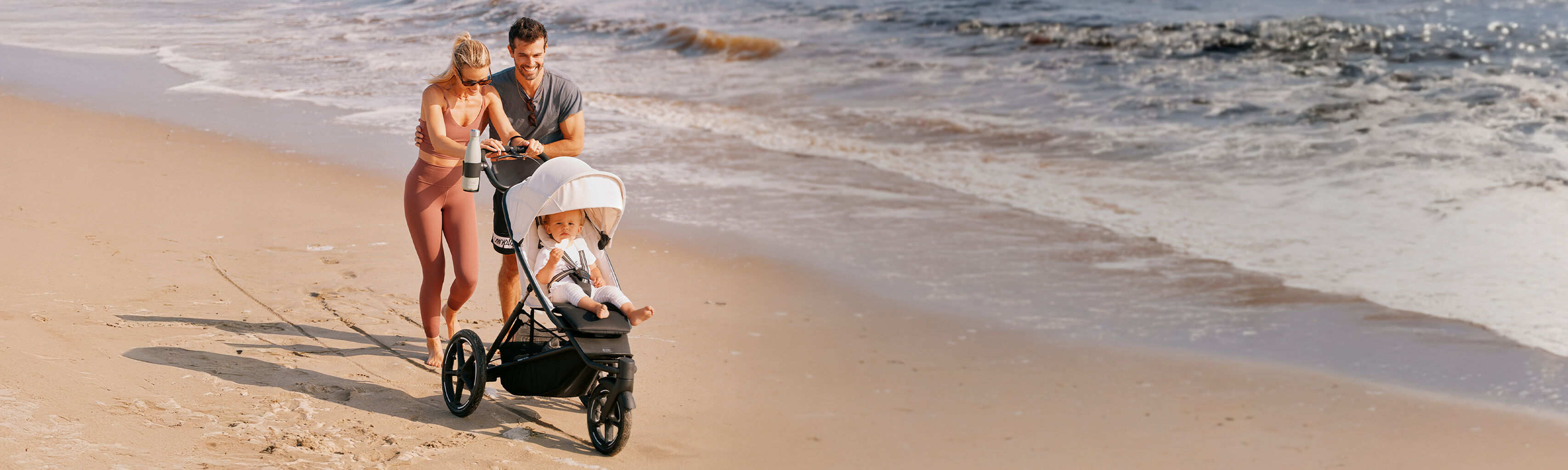 Eine Familie mit einem Kleinkind im Kinderwagen geht am Strand entlang, w&auml;hrend die Wellen sanft im Hintergrund pl&auml;tschern.