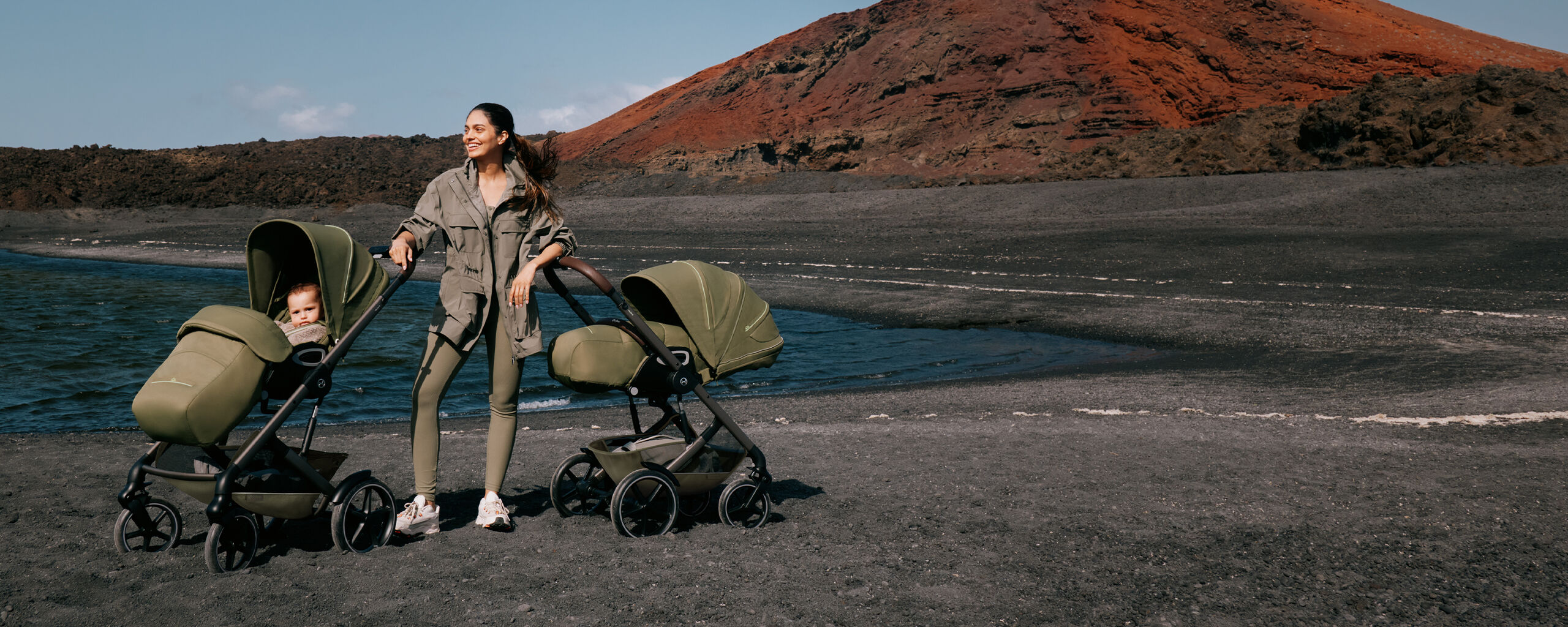 A woman standing on a stony beach with two Talos S Lux strollers, the ocean behind her.